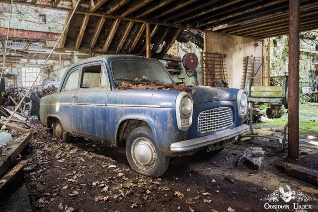 An abandoned, rusty blue 1950s Ford Popular with license plate rests inside a decaying industrial building,