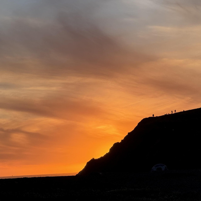 the sark silhouette of the headlands cliff, backlit by a deep orange sunset. a few tiny people can be seen along the edge