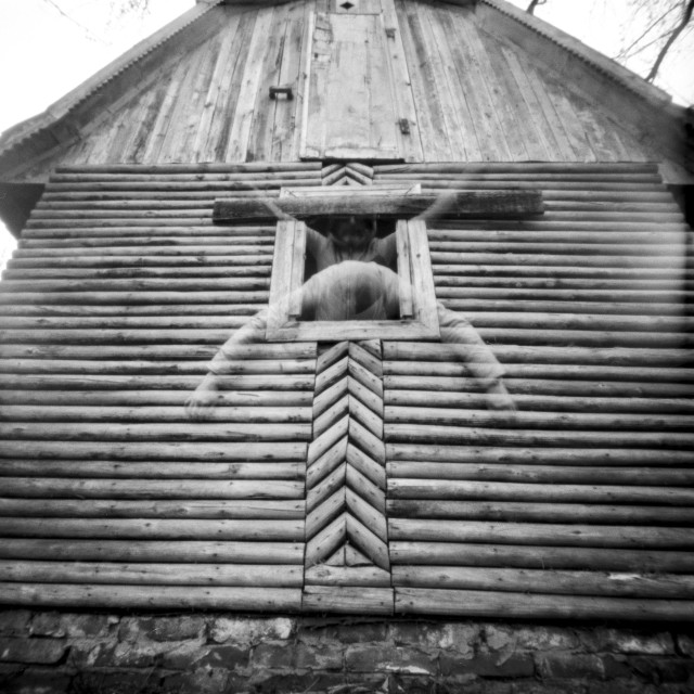 A black-and-white pinhole photograph of a rustic wooden building, seen from a low, symmetrical angle. The vertical planks and geometric patterns of the facade create a strong, almost ritualistic structure, leading the eye toward a small window near the center.

In the window, a human figure appears ghostlike due to the long exposure. The person’s movement is split into two faint positions: in one, they lean downward out of the window with arms extended wide; in the other, they rise upward inside the frame, arms still spread. The overlapping forms create a blurred, spectral presence, as if the body is dissolving or oscillating between two states.

The softness and slight distortion typical of pinhole photography enhance the dreamlike, uncanny atmosphere, turning a simple gesture into something ambiguous—part emergence, part disappearance.