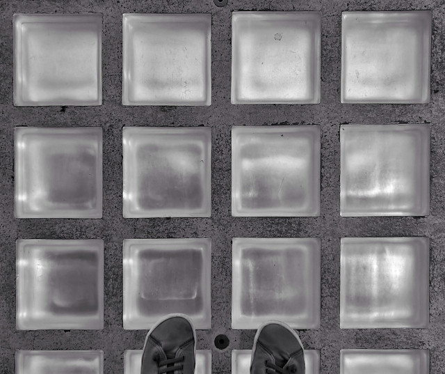 A photo showing part of paving made of square glass bricks, arranged in a grid, lit from below. The tips of a pair of shoes can be seen at the bottom.