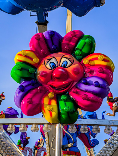A colorful, flower-shaped carousel ride decoration with a smiling face in the center against a clear blue sky. 

The flower's petals are a mix of pink, purple, green, and yellow, and each petal is adorned with small, round lights. The face in the center of the flower has large blue eyes, a red nose, and a wide, friendly smile.

The lighting is bright and sunny, creating a cheerful and festive atmosphere.