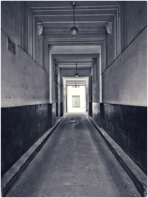 An entranceway to a building is shown and towards the back in the distance it opens up to an area and in that area illuminated in bright sunlight is a door.