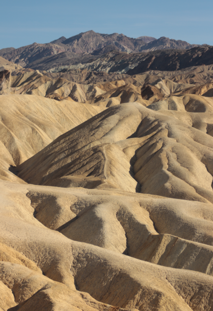 beige and tan hills seem to undulate in massive geological waves as they extend into the distance and rise into a mountain range under a clear blue sky