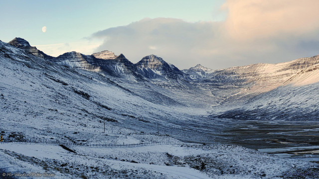 A colour photo of a snow-covered landscape under a partly cloudy sky. The view is of the rounded end of a fjord, looking away from the body of water at a curved glacial semi-bowl of rock. Tall peaks form the crest on the left. The foreground is a road going off to the left and curving round across the lower half of the shot where a river is snaking through on the right. The snowy and icy ground is mostly in blue shadow from the low sunlight from the left, except for the end of the fjord head on the right, which is catching some light. Above the peaks on the left is a gibbous moon.