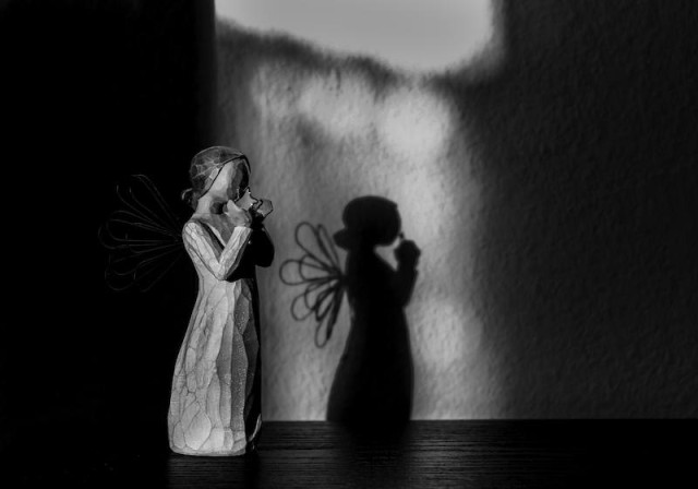 Man sieht auf dem Foto einen geschnitzten Engel, dessen Flügel aus Draht geformt sind. Dieser  Engel wirft einen identischen Schatten an die Wand hinter ihm. Das Foto ist in Schwarzweiß