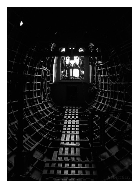 Monochrome image taken from the inside of a vintage plane. The interior is dark with details of the plane's structure just visible. At the far end the cockpit is visible, lit by daylight through the cockpit windows.