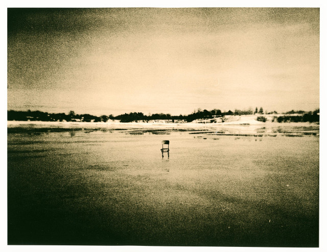 A grainy lith print with a warm, sepia-green tonality shows a vast frozen lake stretching toward a distant, low horizon. In the center of the frame stands a solitary chair, placed directly on the ice—small, fragile, and oddly deliberate against the emptiness.

The shoreline in the distance forms a dark, uneven band of trees and low structures, softened by the lith process into a hazy silhouette. The sky above is luminous yet heavy, its texture mottled with coarse grain that fades from light to deep shadow toward the edges, creating a natural vignette.

The Holga’s character is evident: slight blur, uneven exposure, and a dreamlike softness that dissolves detail into atmosphere. The chair becomes the quiet focal point—a presence without a person, suggesting absence, waiting, or a moment suspended between stillness and disappearance.