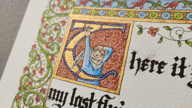 Close up on the letter T and the person within. The inside of the T is painted gold, and the background of the box it's contained in has fine white patterns on dark red.