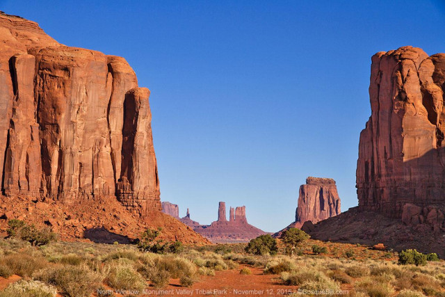 Two massive, sheer sandstone buttes, then a third, frame a darker set of pinnacles in the distance. Blue sky.
©BosqueBill.com
