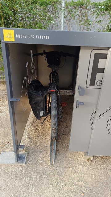 Black bicycle parked in a grey bicycle locker with a black pannier bag on the left side of the rear rack. Sliding door is partially open
