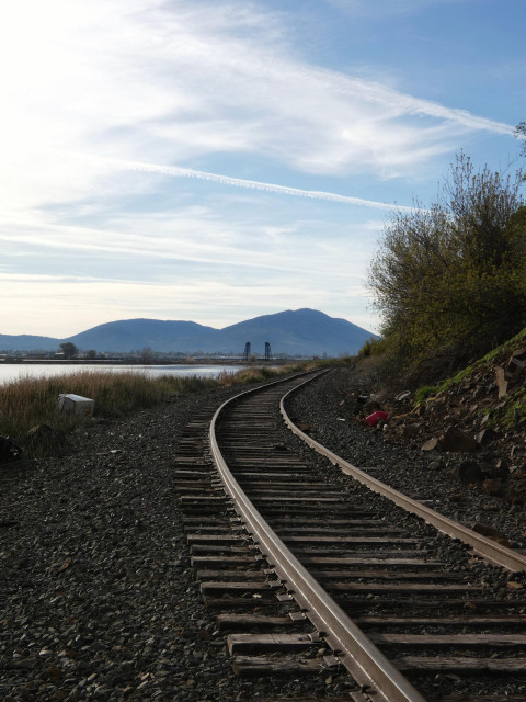 Vertical photo of defunct train tracks curving right around the edge of a rocky foothill, a quiet shimmer of lake reservoir under a wispy morning sky of pale blue, white feathering, and contrails that cut across like tracks themselves. The gravel and tracks on the ground are shadowy and secret, but if you look closely you'll find that humans have tumbled things like old appliances or gas tubs down the hillside, which now have settled there. It's quiet, pretty, and sad.