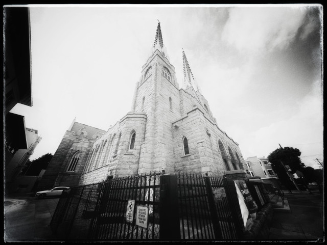 A wide-angle view, looking up from the sidewalk at Saint Paul's Catholic Church in Noe Valley, San Francisco. Monochrome. 

Built between 1897 and 1911, this massive stone church with two tall spires stands tall... but also squats heavily near the southern end of Church Street. It is hard to miss.

Anyway, the church was featured in the 1992 Whoopi Goldberg movie Sister Act. Near the end of the movie, the fictional Pope (who was soft on crime) attended a concert... where Whoopi and her singing nuns sang My Guy (referring informally to God) with great volume and enthusiasm.