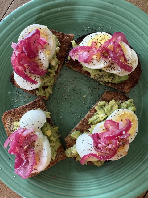 Four pieces of toast on a green plate with green avocado, white egg slices with yellow centers, and bright pink onion garnish on top.