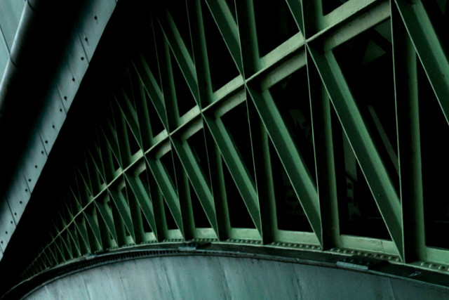 Tilted photo of a section of a green-painted iron arch bridge, the repeating and reflected triangles between the deck and the top of the arch, that swoops across the bottom of the frame, where everything converges at the bottom-left.