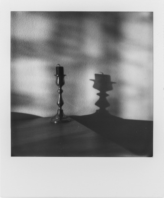 A monochrome Polaroid shows a single candle set in a slender metal candlestick, placed on a flat surface against a plain wall. The candle is unlit, its wick slightly bent. Strong directional light casts a long, sharp shadow across the table and onto the wall, where the shadow of the candlestick appears thicker and somewhat distorted, almost like a different object. Soft, dappled light patterns—resembling reflections or light filtered through leaves—play across the background, contrasting with the stark geometry of the shadow.
