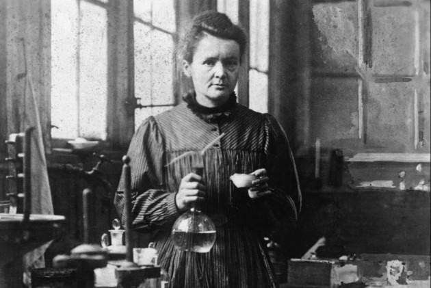 posed photograph of Maria Skłodowska-Curie in her lab holding a volumetric flask. She is a white woman with dark hair in a bun.
