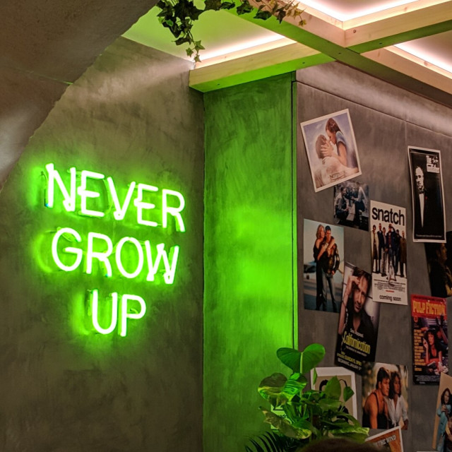 Interior shot of a wall in a trendy cafe featuring a bright green neon "NEVER GROW UP" sign. To the right vintage movie posters are displayed including Snatch, The Godfather, and Pulp Fiction.