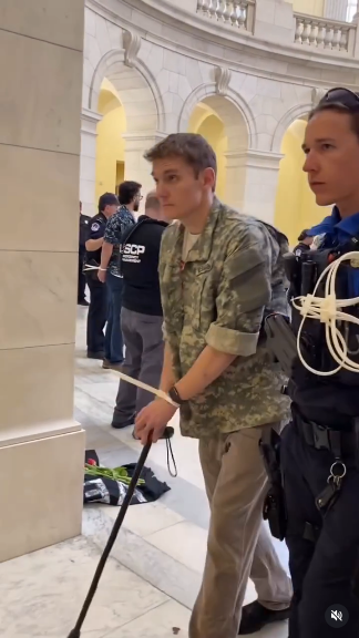 A young man is using a cane for mobility in the Capitol Rotunda. His wrists are zip tied. Next to him is some douchey Capitol police officer with a whole bunch of zip ties strapped to her vest.