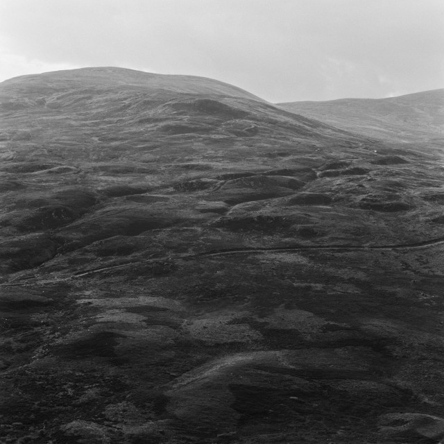 Monochrome photograph of a rolling Highland hillside, its surface textured with peat hags and tussock grass. The hill rises to a broad, rounded summit against a flat grey sky. No human presence, the landscape fills the entire frame.