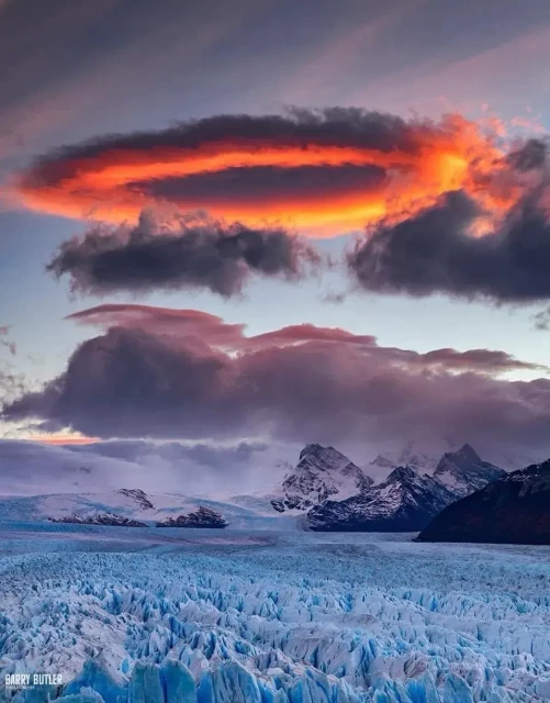 A circular lense shaped cloud in a blue grey sky, but lit by dusk sunset creating a fiery ring in the sky over a blue white Glacier before us.