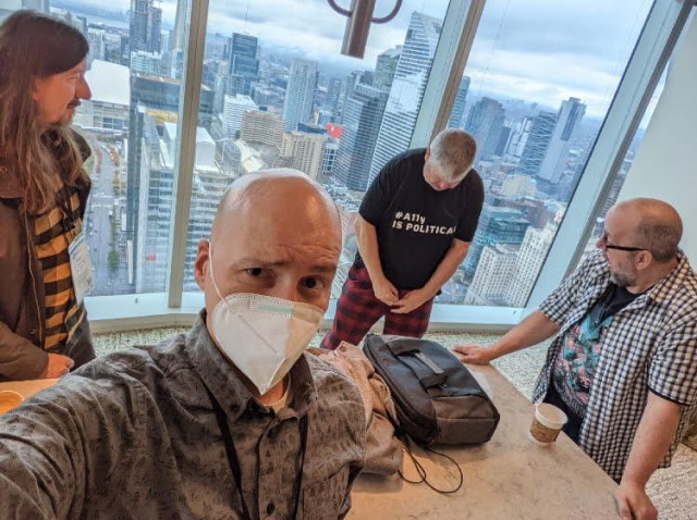 Selfie by Adrian Roselli with me in background attemtping to prove that I was wearing trousers (as they have a zip) rather than Pyjamas. Patrick Lauke are looking on with amazement and disgust at my attempts. There are floor to ceiling windows behind me. The Toronto city skyline can be seen.