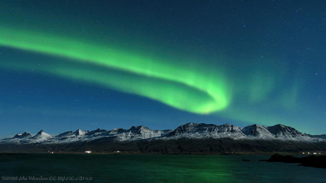 A colour photo of a night sky with lots of stars visible. The foreground is a wide body of water with a range of mountains on the other shore, moonlit from the right. Just above the peaks is a bright green loop of aurora, some places are slightly brighter than others, some parts are quite faint, and a few small flickers of green are visible on the right. The shape is a hook, the upper part brighter than the lower. It's so bright that it's reflected in the water surface.

EXIF: Canon PowerShot G3X, f/2.8, ISO-800, 10 second exposure.