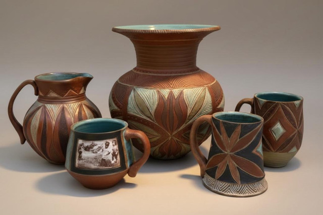 A collection of decorative pottery, including a tall vase, two mugs, a small pitcher, and a bowl featuring intricate geometric patterns. The pieces display a mix of earthy tones and teal accents. One mug features a decal of an old photograph. 