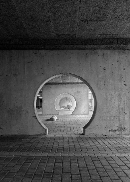 A black-and-white photograph of a series of circular, tunnel-like openings in concrete walls. The walls are part of an parking space below an office building. The floor is paved with rectangular bricks.