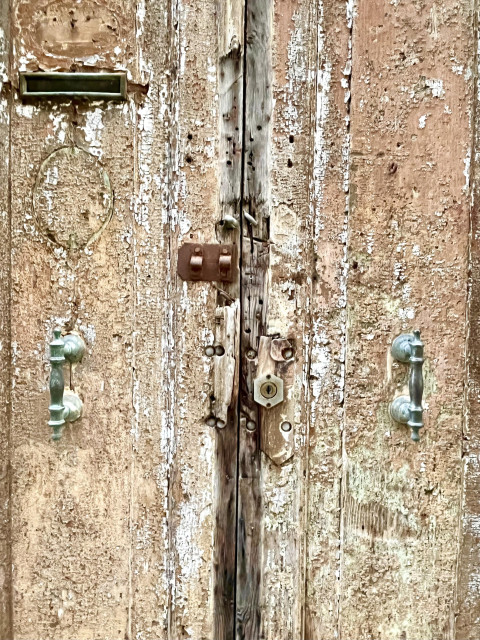Colour photo of the most worn-looking external wooden double door you can possibly imagine. Each side of the door is a solid plank of wood, which might at some point in the distant past have been painted white on top of a rust-red colour, but only a memory of the paint remains as a faint stain on the pockmarked, rough-grained surface. Each side also has a vertical metal handle, rendered verdigris by time and use. There is a narrow metal slot for letters in the top left corner, warped and bent out of shape. The doors were once held shut by a bolt, which has long since disappeared leaving only the rusted metal plate that the bold would have slid into, and holes where the other fixture used to be. It looks more like a stage prop that an enthusiastic designer over-elaborated to the point of parody rather than something which actually exists in the real world.