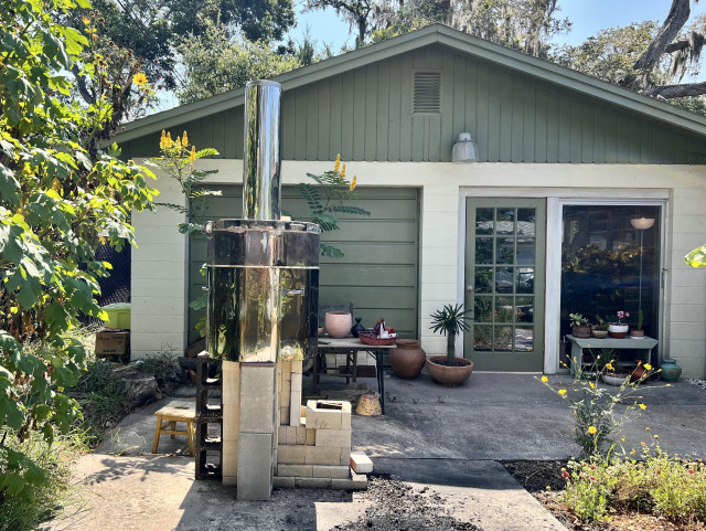 A backyard scene featuring a garage with green trim. In the foreground, there's a kiln with a metal stovepipe sitting on concrete blocks. Surrounding the area are various plants, garden pots, and some outdoor furniture. It’s a sunny day with the light blue sky barely visible behind the garage and trees. 