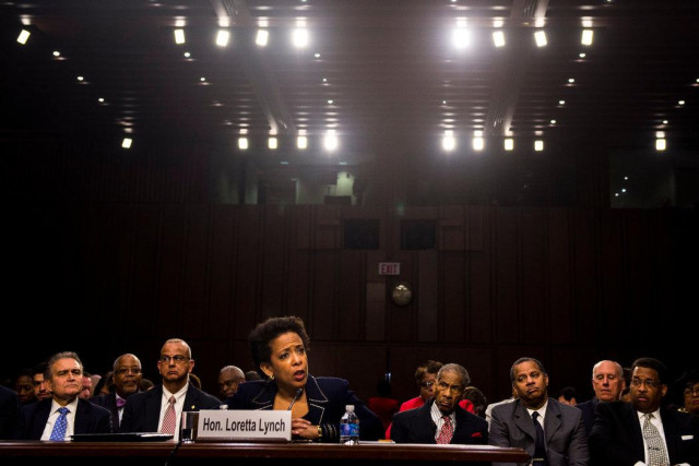 Loretta Lynch at her confirmation hearing: she is a black woman seated at a table whilst rows of men - black and white - sit behind her. the photo is framed to show the number of spotlights she is under.