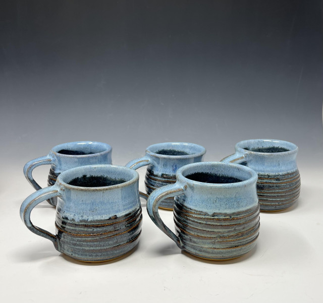Five mugs glazed with two tones of blue: Floating Blue throughout and then a dip of the rims in Lynette's Opal gives bright blue at the top half. Handles are all pointing to the left. The bellies of the mugs have concentric grooved lines that were produced with the sharp tip of a wooden rib while the pots were spinning on the pottery wheel after being formed.