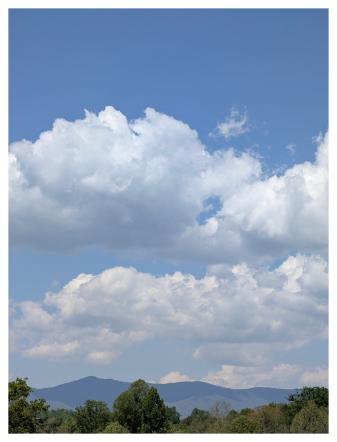 A clear blue sky with large, fluffy white clouds above distant blue mountains. Green trees fill the foreground.