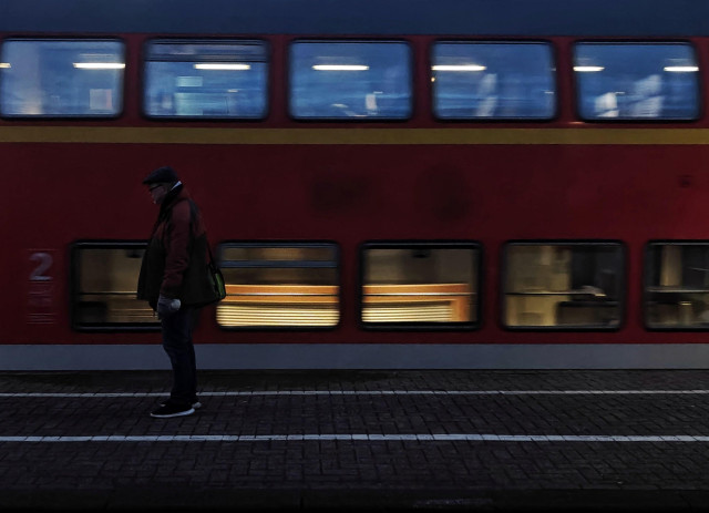 A photo with a person standing on a train platform. Behind the person a motion blurred double-decker train passes. The scene is almost dark.