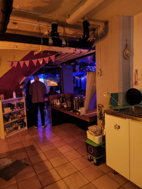 An indoor basement space with beige floor tiles and exposed ceiling pipes. To the left, a person stands near a white shelf; to the right is a small kitchen area with a white cabinet and sink.

​The background transitions into a dimly lit room decorated with blue and purple lights, watermelon banners, and a sign that reads "SOLIDARIA". A wooden counter in the center holds bottles and large metal pots, suggesting a communal or event setting.

