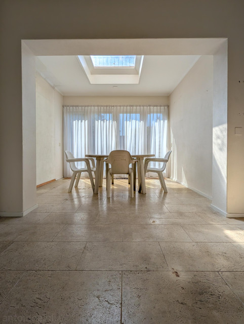 A minimalist indoor space dominated by neutral tones and bright, natural light. The shot is taken from a low angle, highlighting large, light-beige floor tiles with visible texture and dark grout lines.

A thick, white rectangular arch frames the central scene, creating a "room within a room" effect. Large glass doors span the back wall, covered by translucent white sheer curtains that soften the intense sunlight streaming in.

In the center sits a simple, round outdoor-style table surrounded by four white plastic armchairs. 

The scene is almost entirely monochromatic, consisting of various shades of off-white, cream, and beige, which enhances the sense of emptiness and quiet.