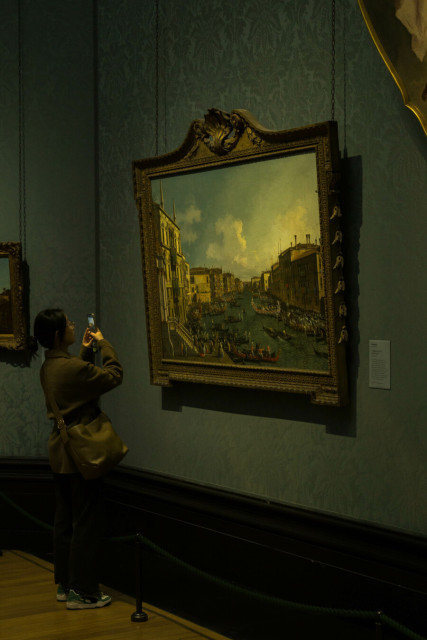 Someone in dark trousers and a taupe jacket and bag leaning slightly back, to the left of the frame, to frame taking a photo on their phone of a Canaletto painting of the Grand Canal in Venice in an ornate gold-coloured frame, hanging against a blue patterned wallpapered wall under yellowish light.