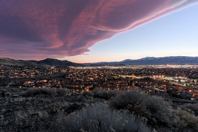 A color landscape photo of a thick line of clouds over a city. It is sunset so the clouds are a reddish color. The thick line of clouds begin in the upper right and extend to center left. From the upper, below the straight line of clouds is a clear wedge shape of light blue sky. Below the sky is a distant mountain range. Under the sky and clouds is a city with all the lights on as it is twilight. In the foreground, where the photography stands, is the top of a hill with a few sagebrush plants.