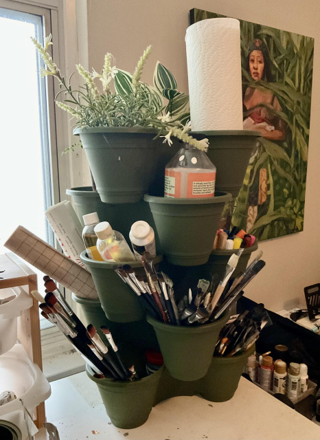 Green stacking planters holding paintbrushes, rolled paper, paper towel, art mediums & fake plants. (Real ones maybe to come later!) sitting on desk. One of my paintings & part of a window on either side. 
