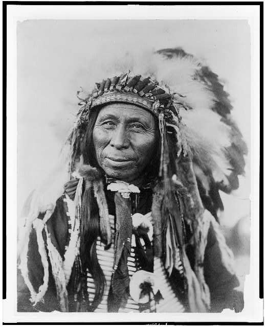 This is a black-and-white head-and-shoulders photograph of Black Thunder, a Dakota (Sioux) man, who is facing slightly left. He is wearing a feathered headdress with beaded banding and long braids, along with layered clothing that includes patterned fabric and a circular pendant at his chest. His expression is serene and steady, with a slight smile.  The background is white and plain, drawing the focus to his face and regalia.