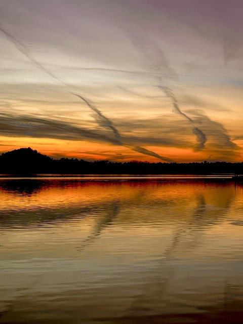 A sunset over a lake that is red, yellow, and orange at the horizon with light blue and purple up top. Wispy threads of dark cloud strands stand out against the color. It is all reflected in the water surface.