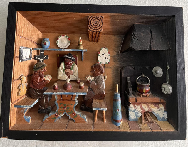 A carved scene (possibly eastern European based on the details) of three men playing cards inside a small cabin. There is a large cross, fiddle and knickknacks hanging on the walls and a large cookstove in the corner. A pot simmers on the wood stove and a blue butter churn sits nearby. The men have very detailed faces with lots of character.
A large timber beam holds up the roof. 
