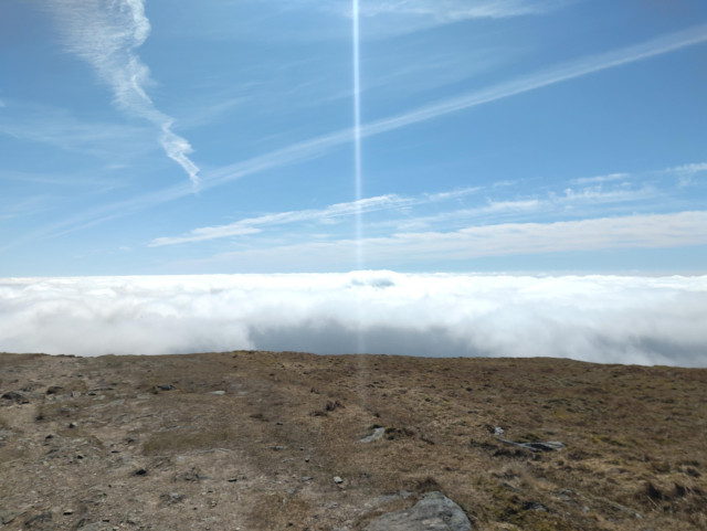 Photo of a stony / mossy landscape with a view over whire, cotton-line clouds seen from above and a bright blue sky above them