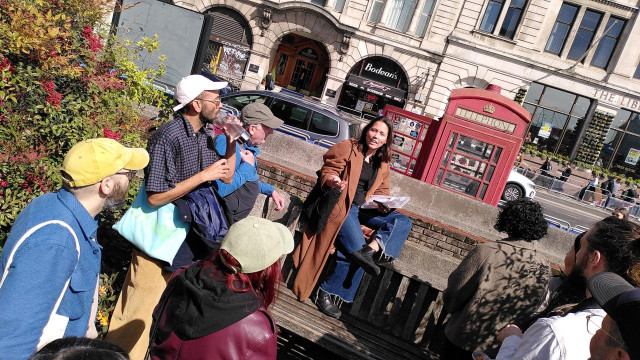 Anne perched on a wall next to a red antique London telephone booth surrounded by standing participants of the Internet Infrastructure Walk in the streets of London.