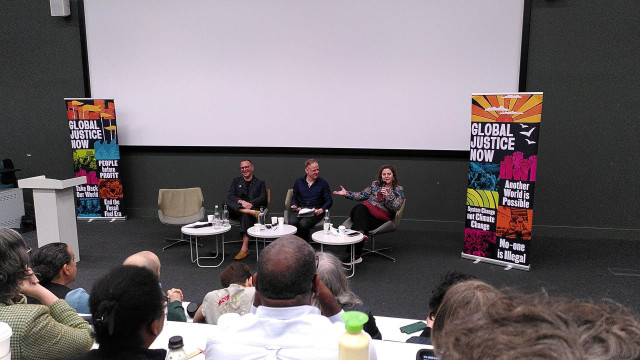 Cory Doctorow and Sofia Scasserra in conversation chaired by
Nick Dearden. Three people sitting on chairs with microphone in the front of an auditorium with a banner for the Resisting Big Tech Empires event projected on a screen.