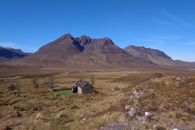 A bothy, a single story stone cottage, sits in the heather. Behind mountains rise up to the blue sky