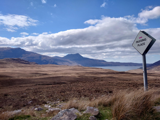 A white diamond "passing place" sign. Beyond it, a vast desolate landscape of heather leads away to a loch and mountains under a blue sky