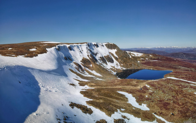 An aerial view of a loch with cliffs covered in snow and a blue sky above. The Stuic and Loch nan Eun. Cairngorms NP, Scotland