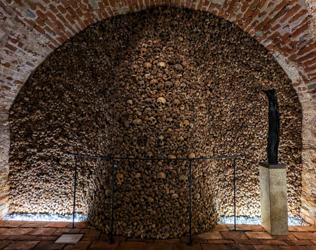 Interior view of the Brno Ossuary, located beneath the Church of St James. The image showcases a large arched chamber constructed from red brick, where the walls and ceiling are meticulously covered with hundreds of human skulls and bones. The bones are arranged in dense, orderly patterns, creating a striking and solemn mosaic that fills the entire visible space within the arch. A simple metal railing runs along the base of the wall, likely serving as a protective barrier for the remains and a guide for visitors. The floor is made of stone tiles, and in the foreground stands a stone pillar featuring a sculpture titled 'Soul,' adding an artistic and contemplative element.