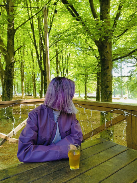 A picture of me with purple hair and purple leather jacket sitting at wooden table in the forest, looking to the side. A glass of cider on the table 